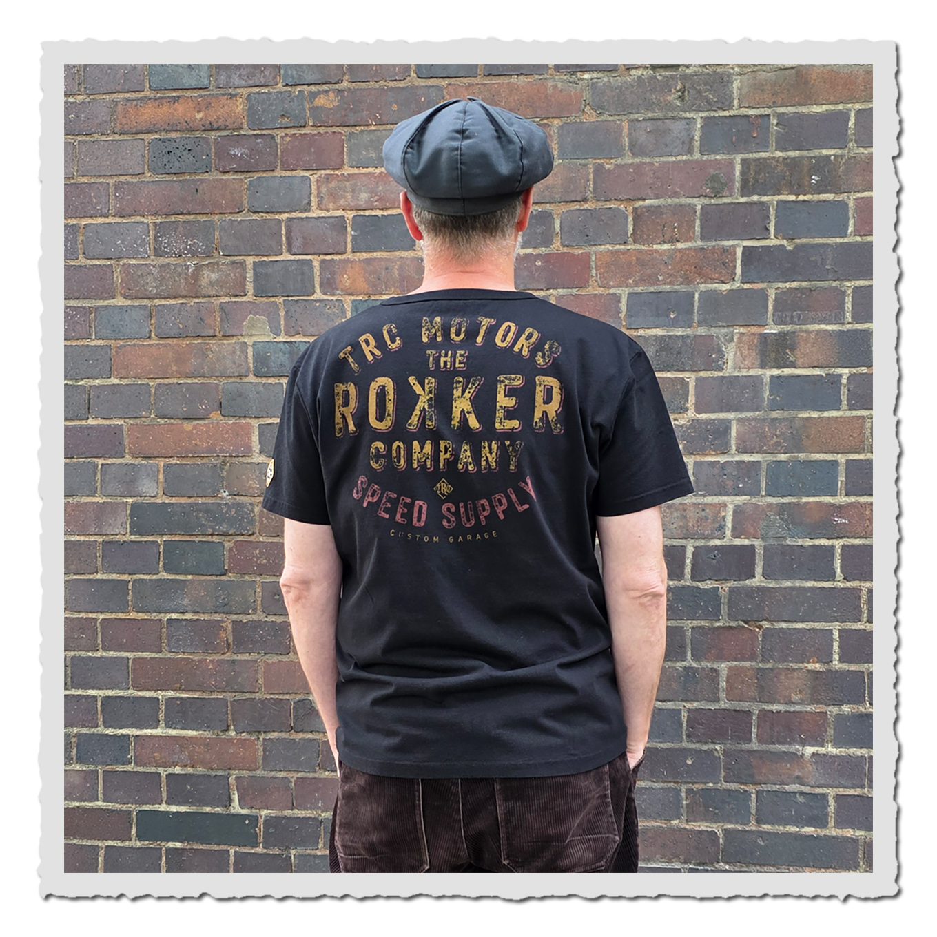 Person trägt schwarzes T-Shirt von hinten mit großem Schriftprint vor braun-grauer Backsteinwand.